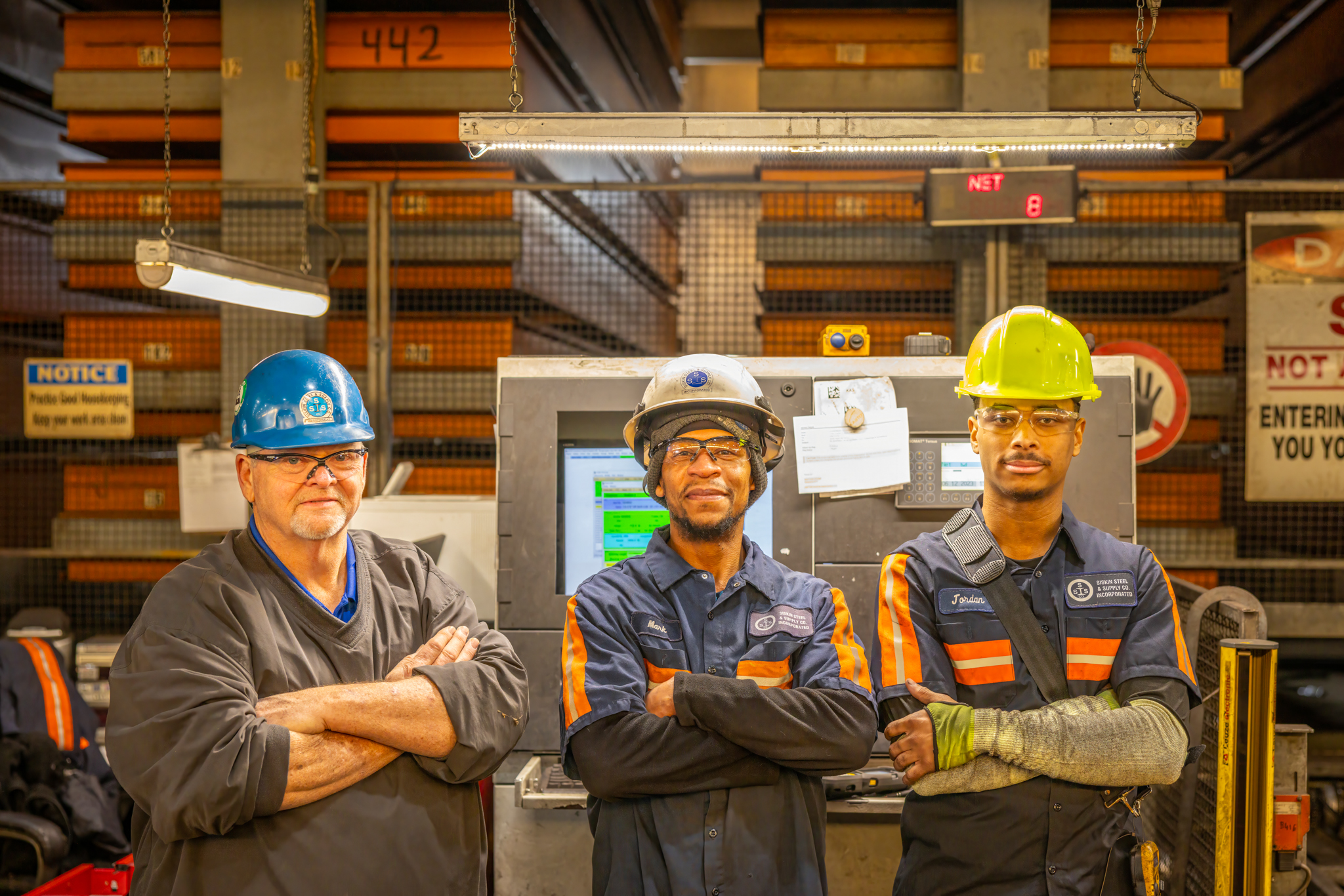 Three men standing side by side while crossing their arms in an industrial warehouse setting with a large computer screen and storage system behind them.