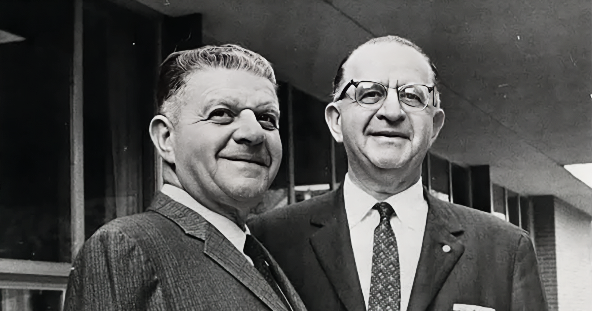 Two men in suits looking up and smiling with a brick office building in the background