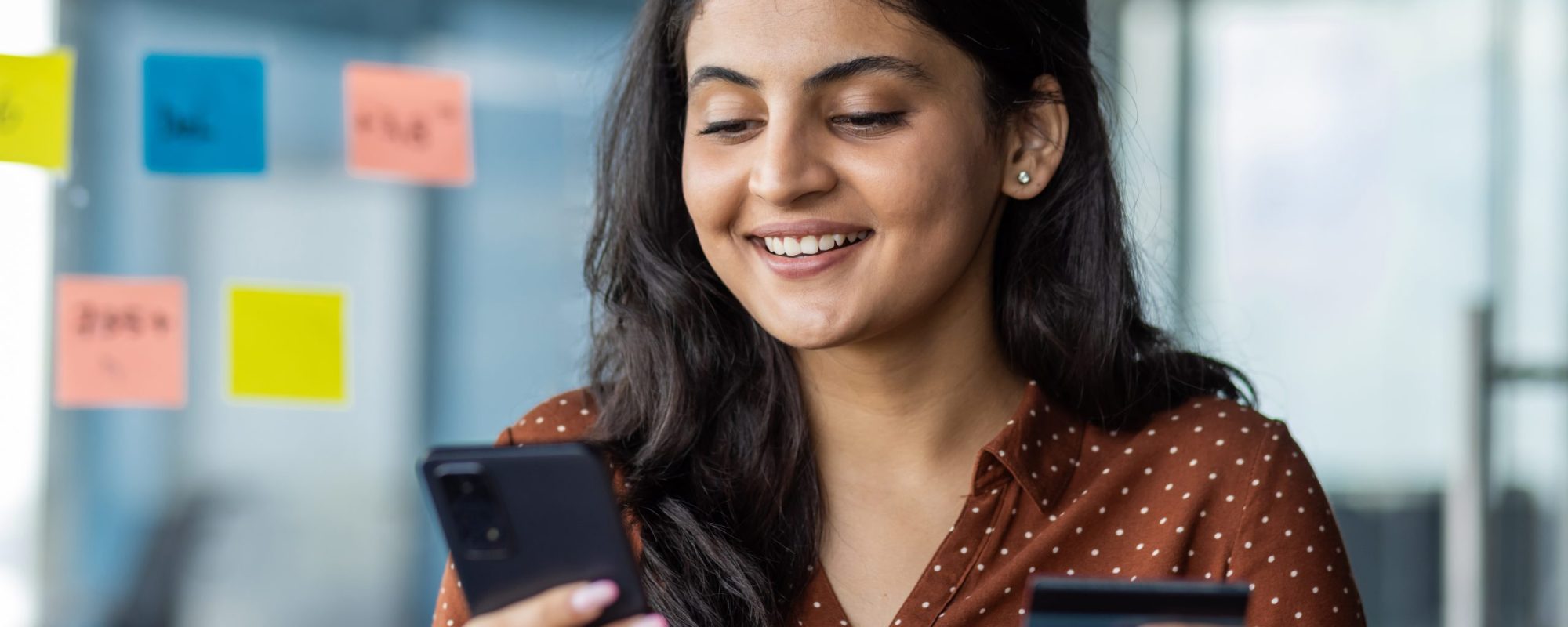 Young woman holding a smart phone in her left hand and a cred card in her right hand inside of an office with glass walls.